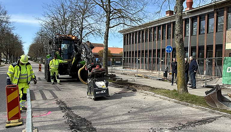 Två män i varselkläder arbetar på en väg med en grävmaskin.